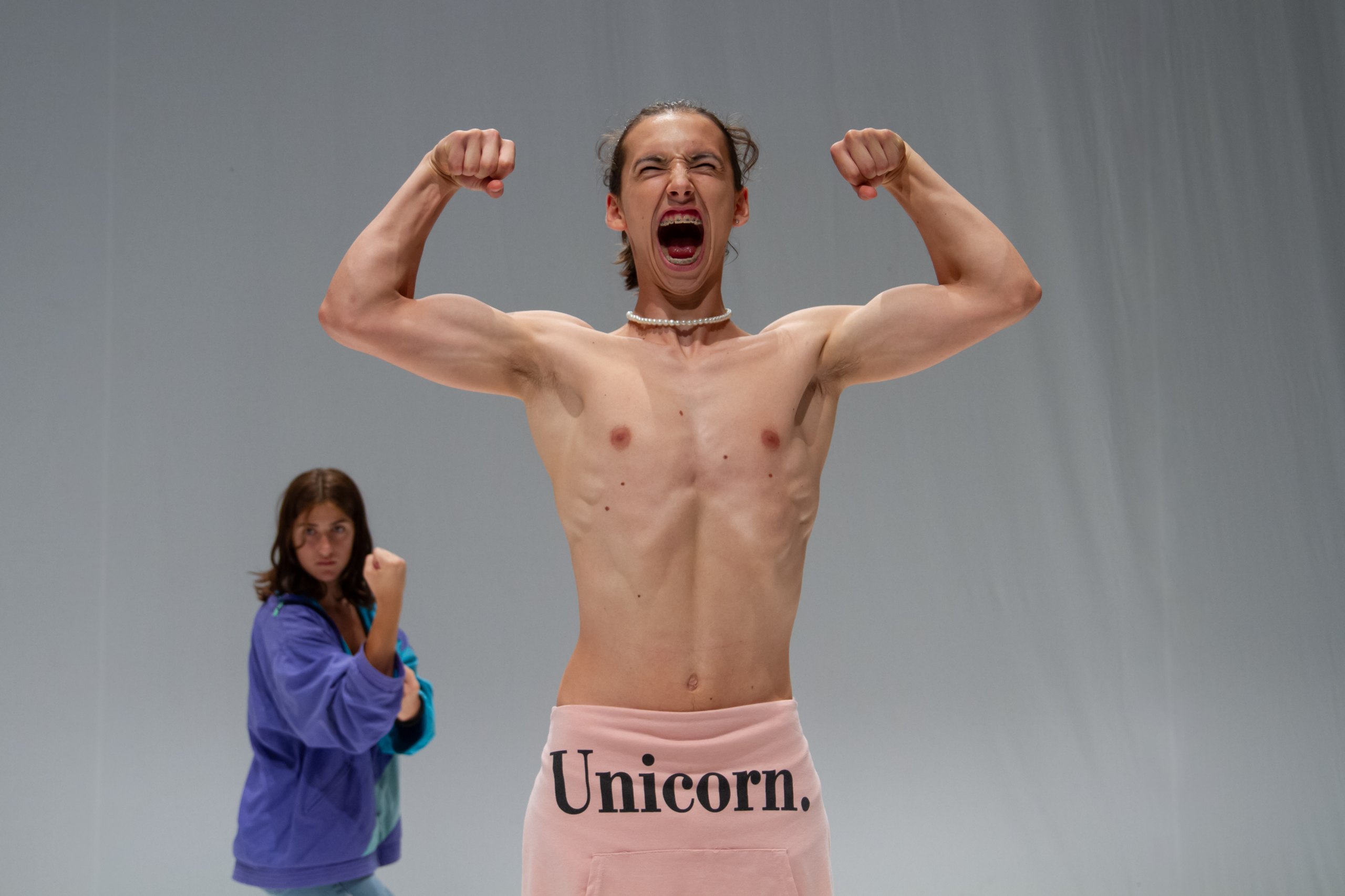 Performer a torso nudo al centro della scena, con le braccia flesse e la bocca aperta; sullo sfondo un'altra performer in posa con i pugni alzati.