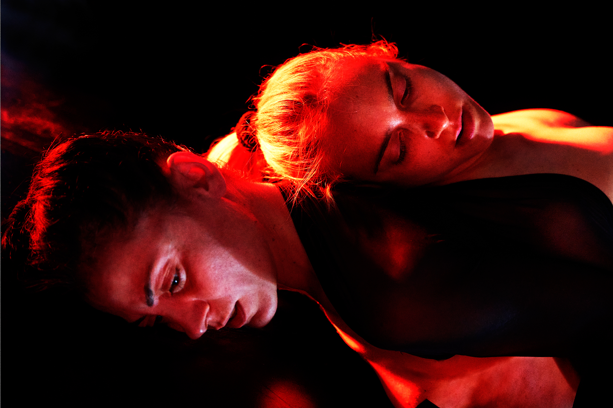 Immagine: due performer, vestite di nero, sono distese e intrecciati su uno sfondo buio. Una luce rossa intensa illumina i loro volti: una persona guarda di lato, l’altra tiene gli occhi chiusi con la testa appoggiata sul suo corpo. Immagine di Fear of the Dark di Cult of Magic.